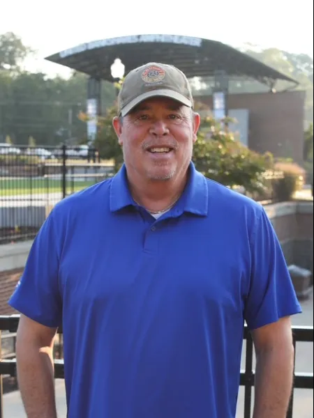 Smiling man in a blue polo shirt and cap standing outdoors near a sports stadium entrance during daytime