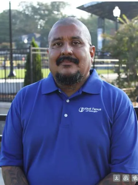Man in a blue First Fence of Georgia polo shirt standing outdoors near a fence and trees.