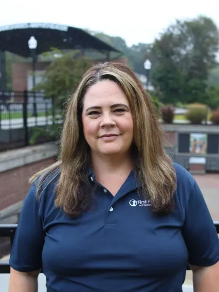 Woman with light brown hair wearing a navy blue First Flight polo shirt outdoors near trees and fencing.