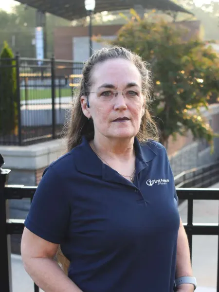 Middle-aged woman wearing glasses and navy blue polo shirt with logo standing outdoors by black fence.