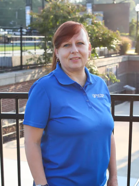 Middle-aged woman in a blue polo shirt standing outdoors in front of a black metal fence with greenery in the background