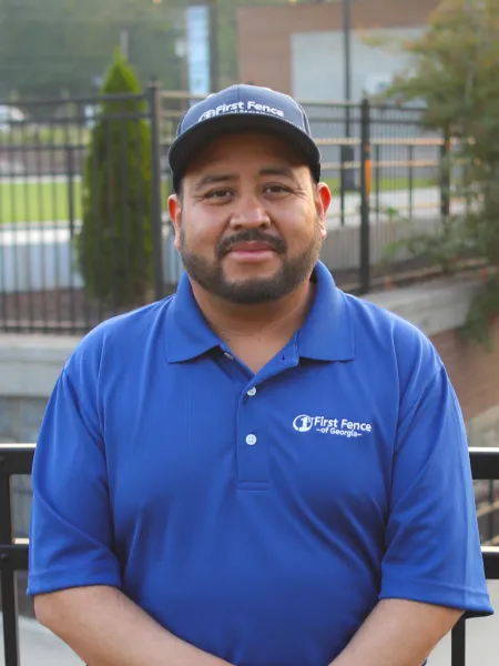 Man wearing a blue First Fence of Georgia shirt and cap standing outdoors by a fence.