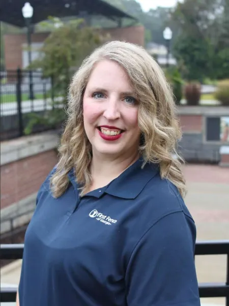 Smiling woman with curly blonde hair wearing a navy blue First Fence polo shirt outdoors.