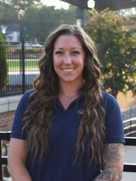 Smiling woman with long wavy hair and tattooed arm wearing a navy blue polo shirt outdoors.