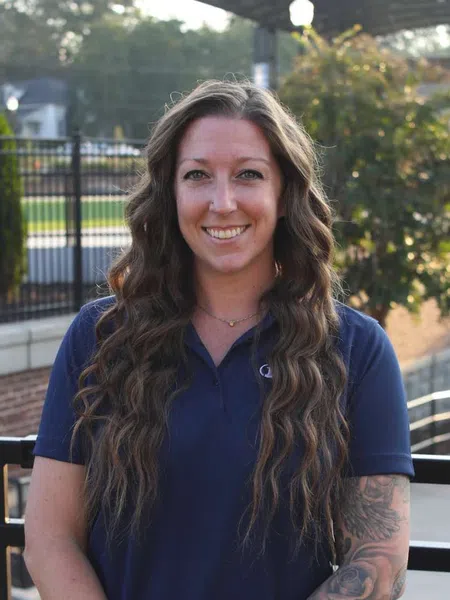 Smiling woman with long wavy hair and tattooed arm wearing a navy blue polo shirt outdoors.