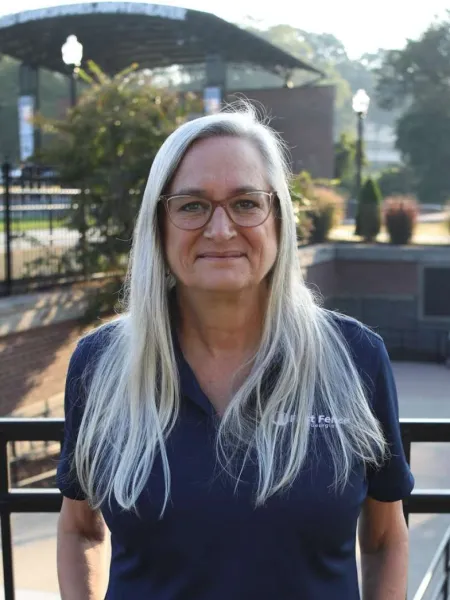 Mature woman with long gray hair and glasses wearing navy blue polo outdoors in daylight.