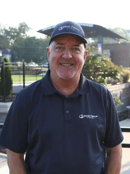 Smiling man wearing a First Fence of Georgia navy cap and polo shirt outdoors near a black fence.