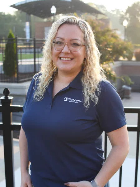 Smiling woman with curly blonde hair wearing glasses and a navy First Fence of Georgia polo shirt outdoors.
