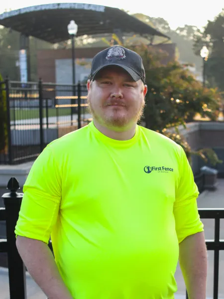Man wearing bright yellow First Fence of Georgia shirt and black cap standing outdoors near black fence.