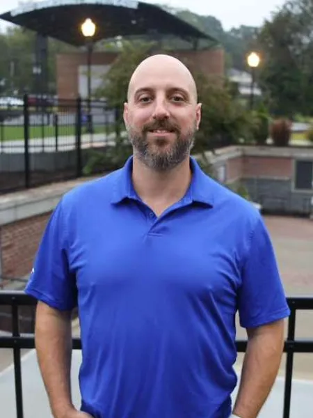 Bald man with beard wearing blue polo shirt and smartwatch standing outdoors with hands in pockets.