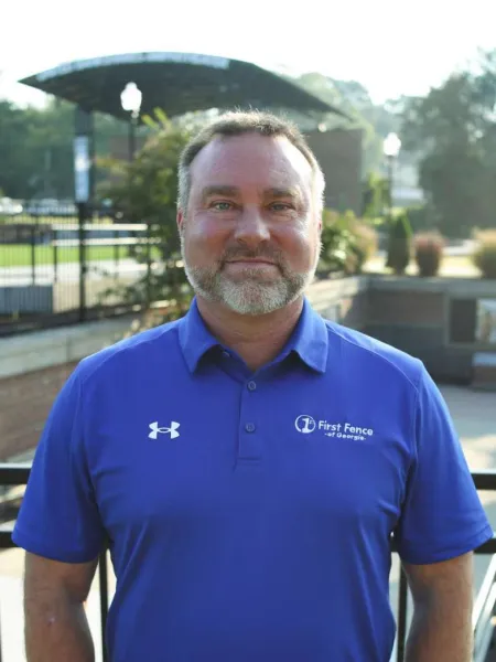 Man wearing a blue First Fence of Georgia polo shirt with Under Armour logo, standing outdoors in a park setting.