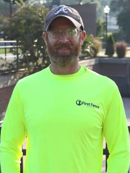Bearded man wearing glasses, a gray baseball cap, and a bright yellow First Fence of Georgia shirt outdoors.
