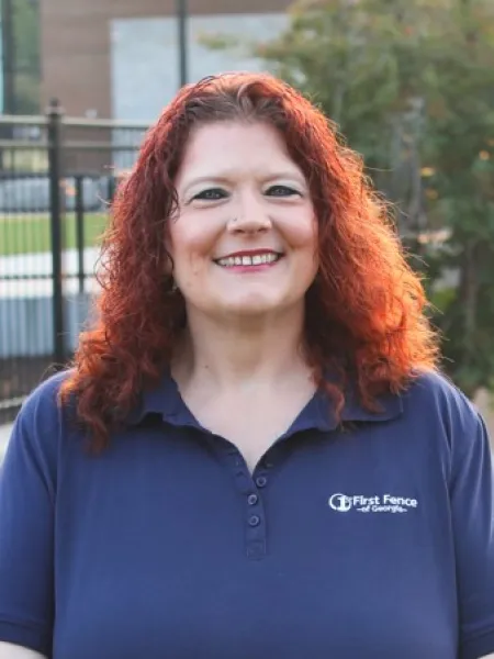 Smiling woman with curly red hair wearing a navy First Fence polo shirt outdoors by a fence.