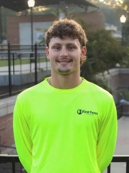 Smiling man wearing a bright yellow First Fence long-sleeve shirt standing outdoors near a fence.