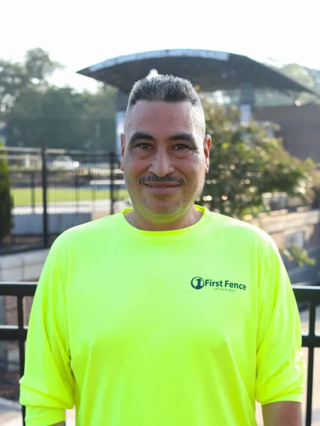 Man wearing a bright yellow First Fence of Georgia shirt smiling outdoors with fencing and trees in the background