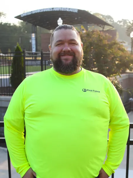 Smiling man with beard wearing bright neon yellow First Fence shirt standing outdoors near amphitheater.