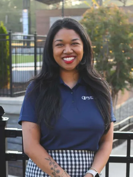 Smiling woman with long black hair wearing a navy polo and checkered skirt standing outdoors with tattoo on arm