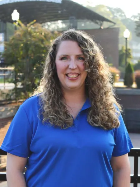 Smiling woman with curly hair wearing a blue polo shirt standing outdoors in a park on a sunny day