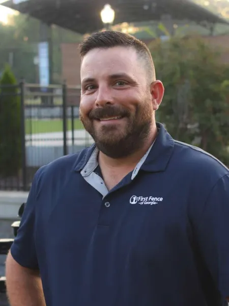 Smiling man with beard wearing a navy blue First Fence of Georgia polo shirt outdoors near a fence.