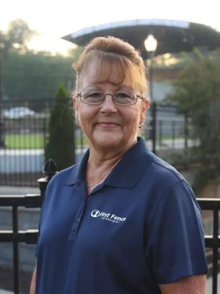 Smiling woman wearing glasses and navy blue First Fence of Georgia polo shirt standing outdoors near a fence.