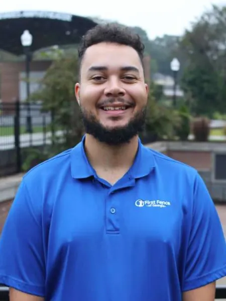 Smiling man wearing a blue First Fence of Georgia polo shirt outdoors near a fenced area and greenery.