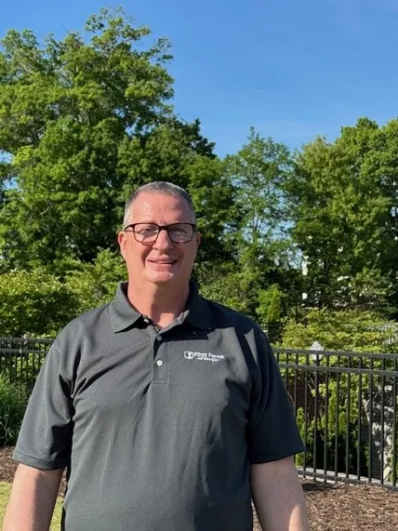 Man wearing glasses and a gray polo shirt standing outdoors with green trees and black metal fence in the background.