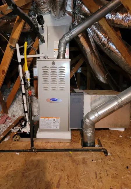 Residential furnace and ductwork installed in an attic with wooden beams and insulation visible.