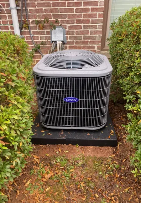Outdoor Carrier air conditioning unit installed on a black platform between green bushes near a brick house.
