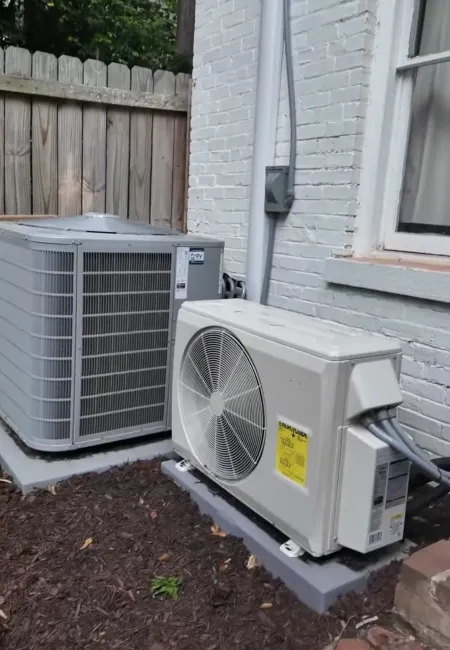 Two outdoor HVAC units installed on concrete pads beside a white brick house and wooden fence.