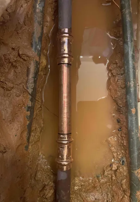 Copper pipe fitting installed in muddy ground with standing water around it during plumbing work.