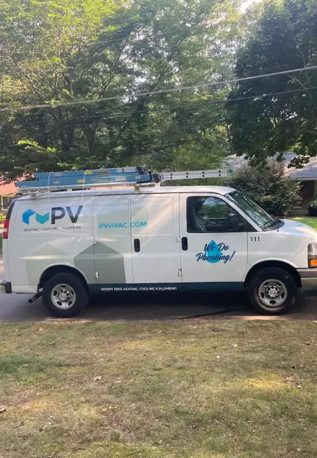 White PV HVAC plumbing service van parked on suburban street with ladders on top and trees in background