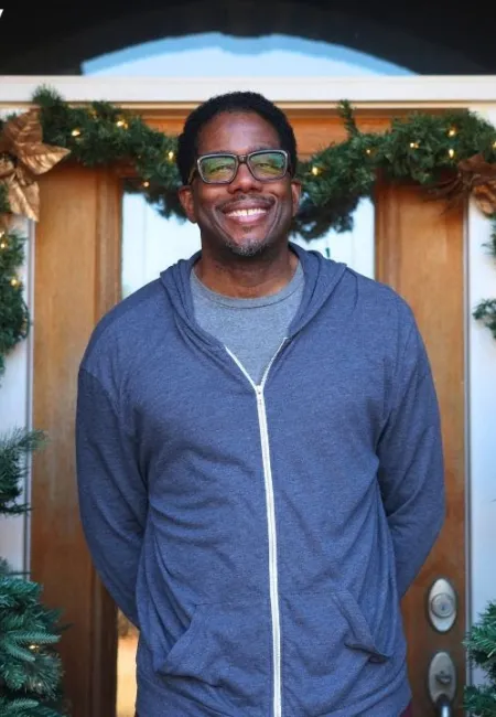 Smiling man in glasses and blue hoodie standing between two decorated Christmas trees in front of a wooden door
