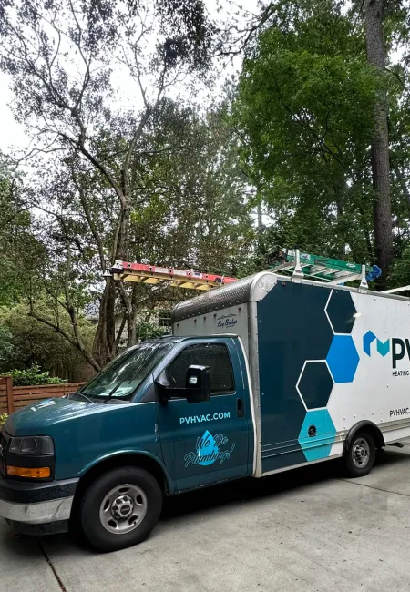 Blue and white PV Heating & Air plumbing service van parked in a wooded residential driveway with ladders on top.