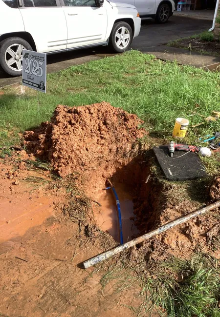 Open muddy trench with blue pipe, tools, and a congratulatory 2025 sign near parked white cars on grass.