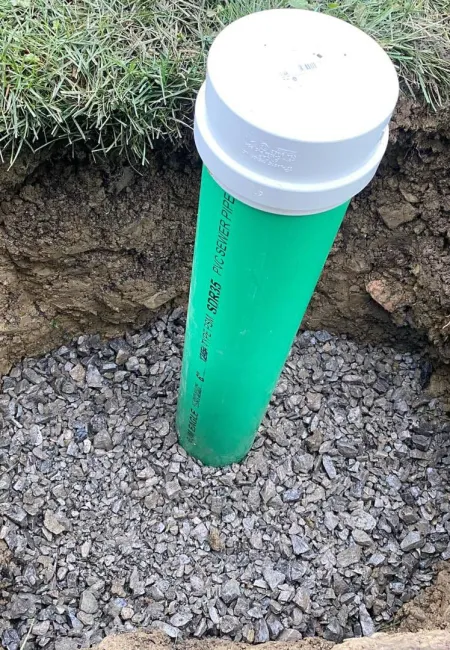 Green PVC pipe with white cap installed in gravel-filled hole surrounded by grass in yard