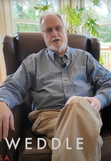 Older man wearing glasses and a checkered shirt sitting in a leather armchair by a window with plants and a lamp nearby