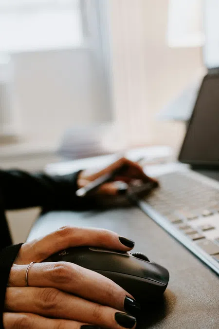 Close-up of hands using a wireless mouse and typing on a laptop keyboard in a bright office.