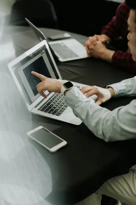 Two people collaborating over laptops and smartphone, one pointing at a laptop screen during discussion.