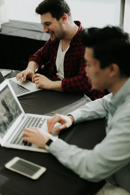 Two men collaborating and smiling while working on laptops at a table with a smartphone nearby