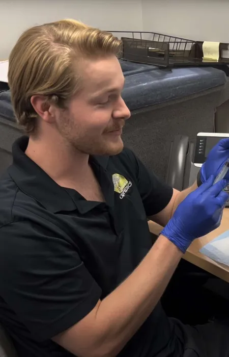 Smiling man wearing blue gloves examining a small vial at a cluttered desk with medical supplies and tools.