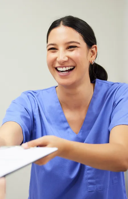 Smiling healthcare professional in blue scrubs handing a clipboard to a patient in a medical setting.