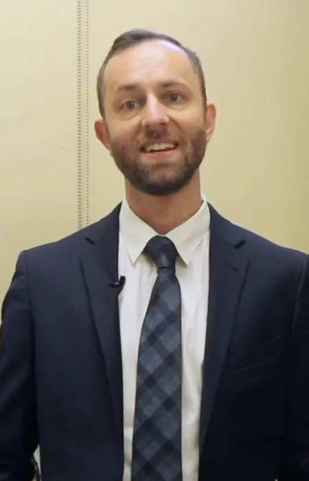 Man in suit standing in medical office with X-ray images displayed on a monitor behind him