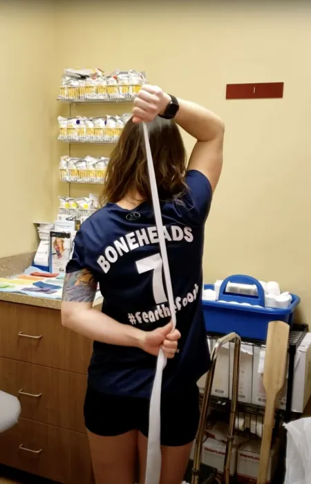 Person in a Boneheads jersey performing shoulder stretch with a band in a medical or therapy room.