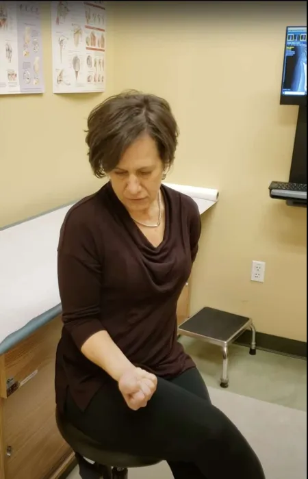 Woman sitting on a stool in a medical exam room testing arm strength with a clenched fist.