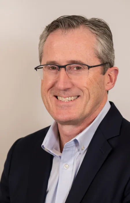 Smiling middle-aged man wearing glasses, a light blue shirt, and a dark blazer against a plain background