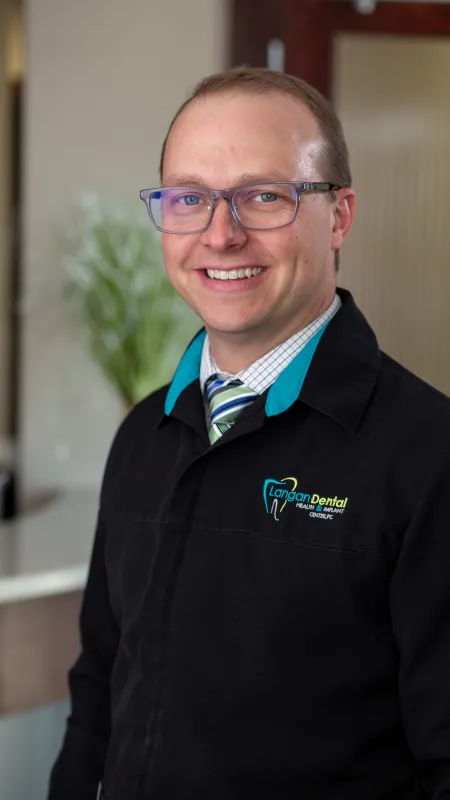 Smiling male dental professional wearing glasses and Langan Dental jacket in modern office setting.