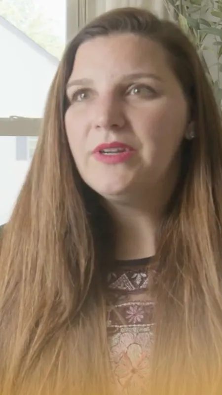 Woman with long brown hair speaking indoors next to a window with a white house outside