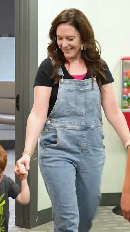 Woman in denim overalls holding a young boy's hand walking in an office with another woman in black shirt