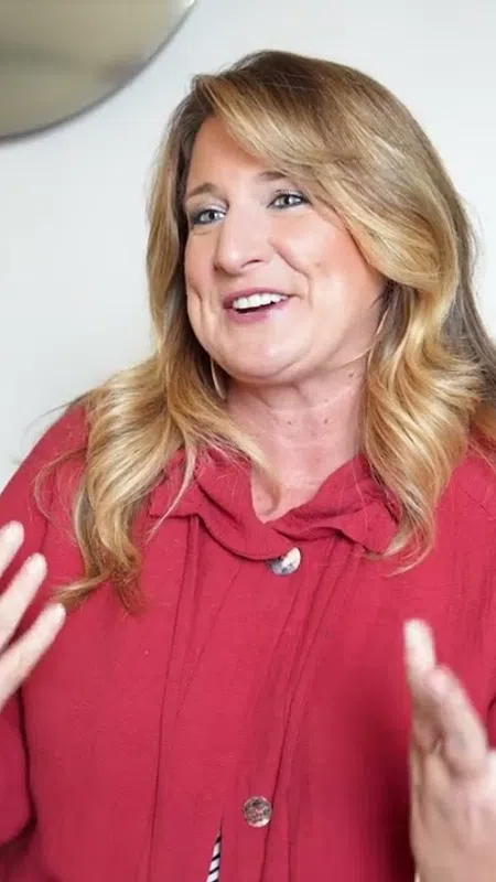 Smiling woman with blonde hair wearing a red sweater gestures while sitting in a modern living room with a gray chair.