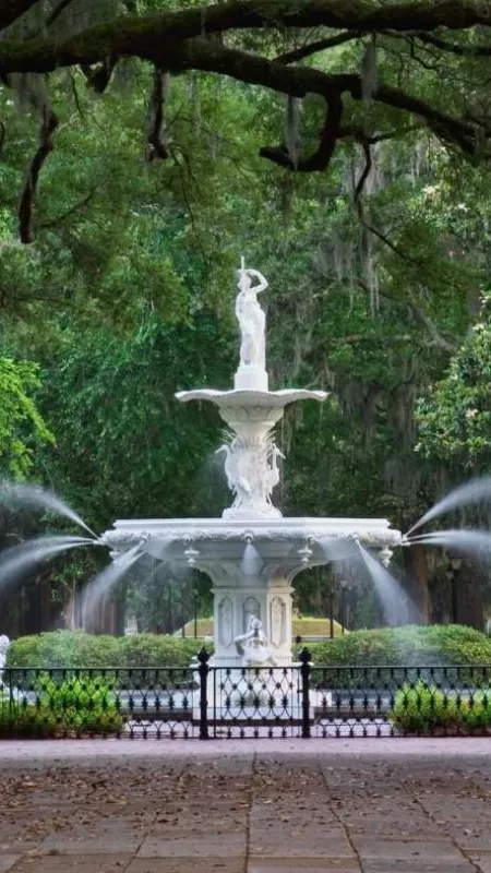 Ornate white fountain with statues in lush green park surrounded by large trees and hanging moss